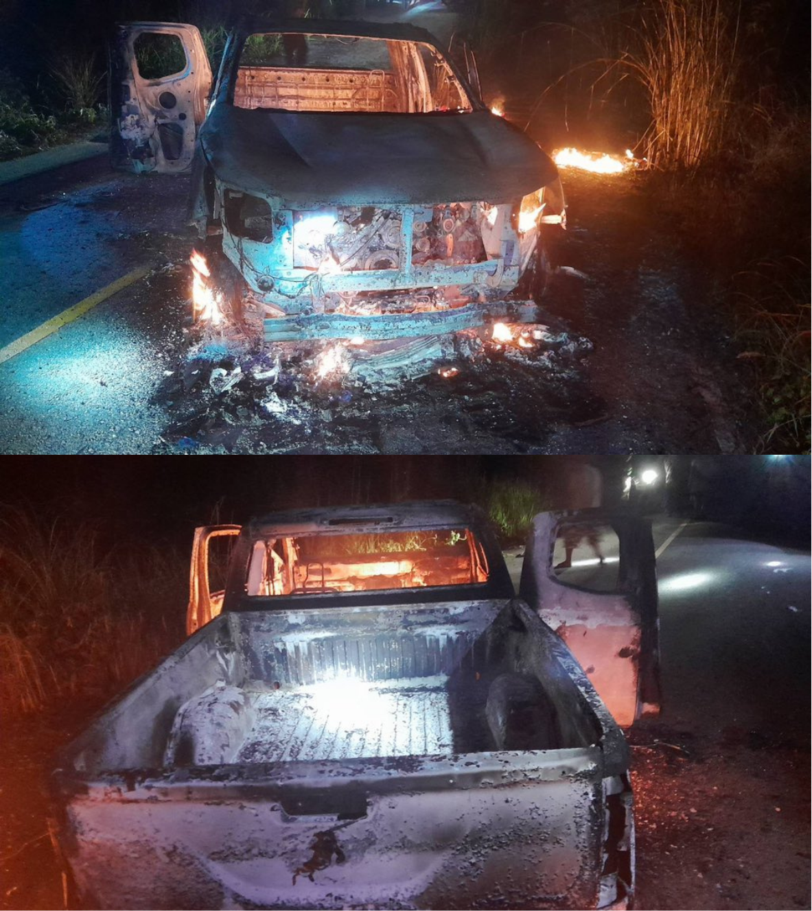 A burned-out pickup truck at night on a roadside, flames still visible in the engine bay and in the grass beside it.