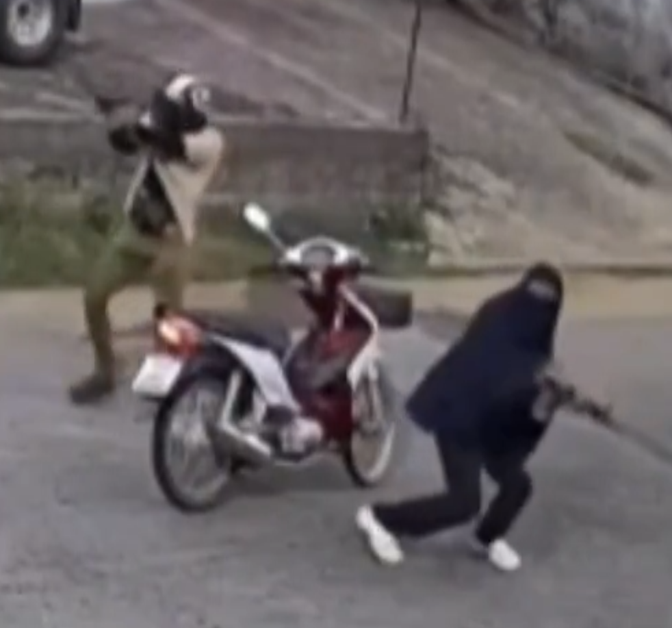 Enlarged CCTV frame showing two people stepping away from a parked red-and-white scooter, one in a full-body black garment carrying a rifle.